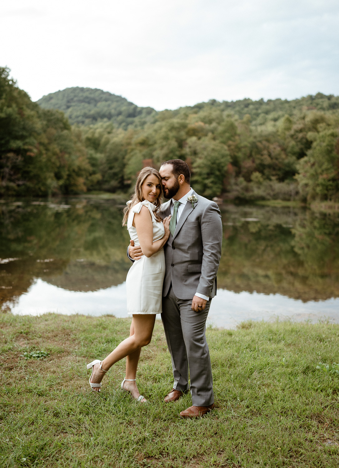 Bride and groom nuzzle in close in front of a mountain lake on their Asheville elopement day